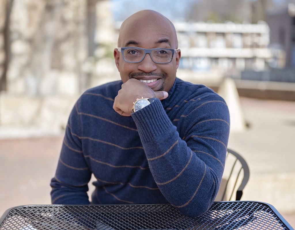 A portrait photo of Kwame Alexander, a Black American man, sitting with his hand to his chin and grinning at the camera.