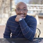 A portrait photo of Kwame Alexander, a Black American man, sitting with his hand to his chin and grinning at the camera.