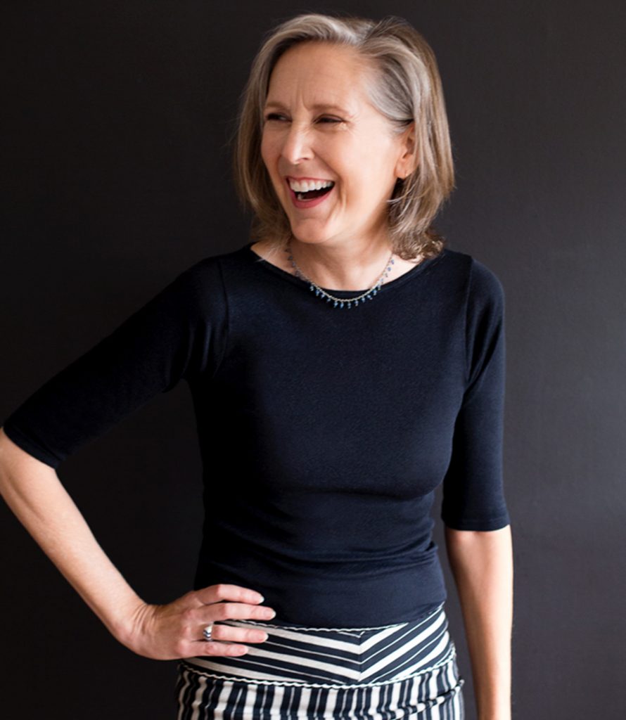 A photo of Mary Roach laughing at something to the left of the camera, with her hand on her hip.
